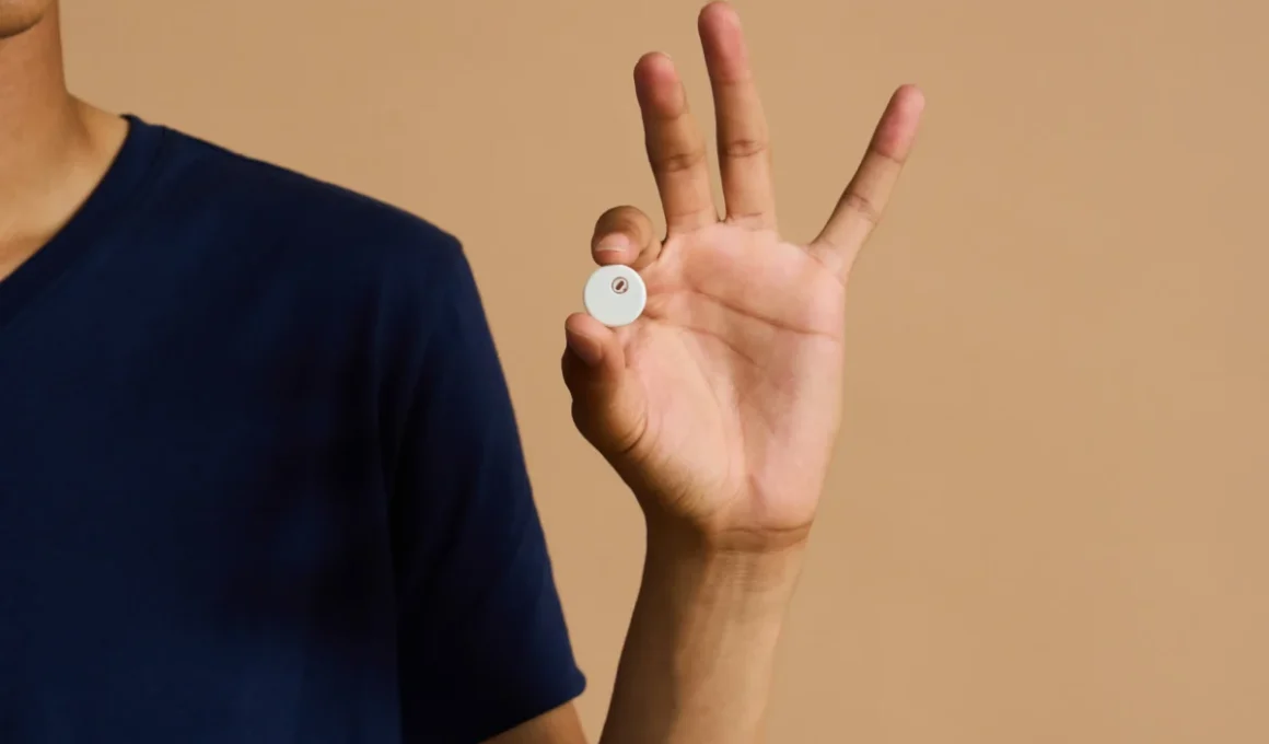A hand holding a small round white sensor about the size of a coin, against a tan background.