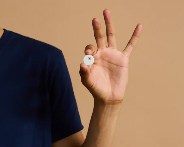 A hand holding a small round white sensor about the size of a coin, against a tan background.