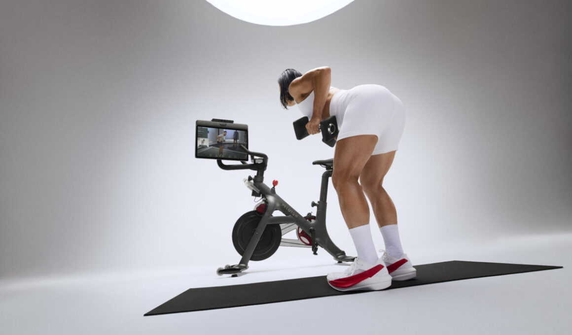 A Peloton Bike on a pale studio backdrop, with an athlete mid-air beside it, one leg driven up and both arms raised.