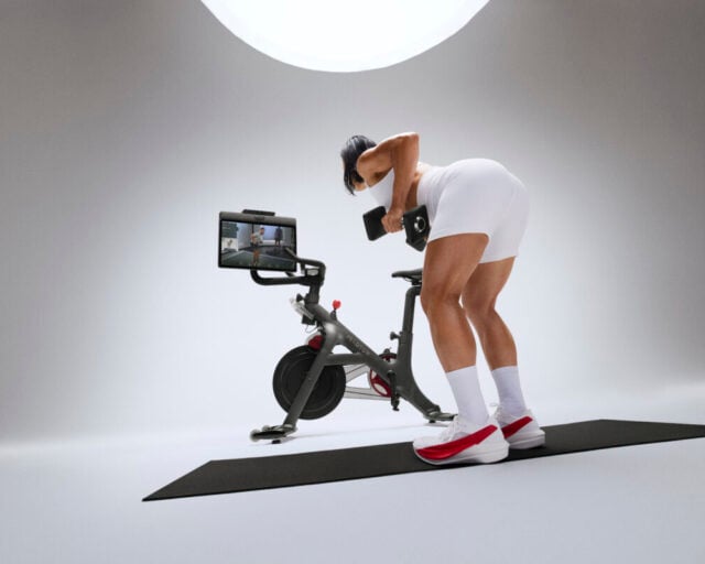A Peloton Bike on a pale studio backdrop, with an athlete mid-air beside it, one leg driven up and both arms raised.