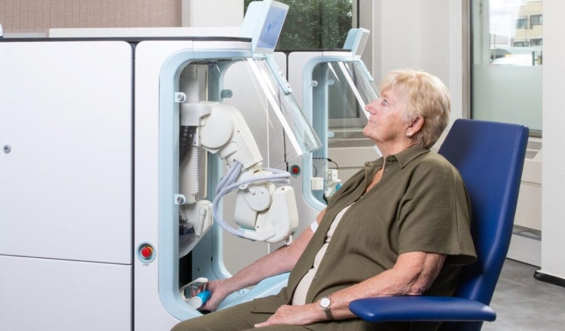 A seated patient with one arm inside the Aletta robotic blood collection device in a clinical room, with a second unit visible behind.