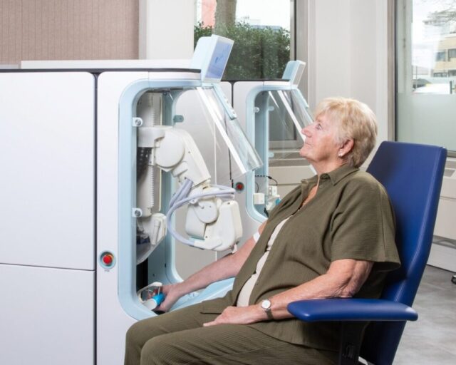 A seated patient with one arm inside the Aletta robotic blood collection device in a clinical room, with a second unit visible behind.