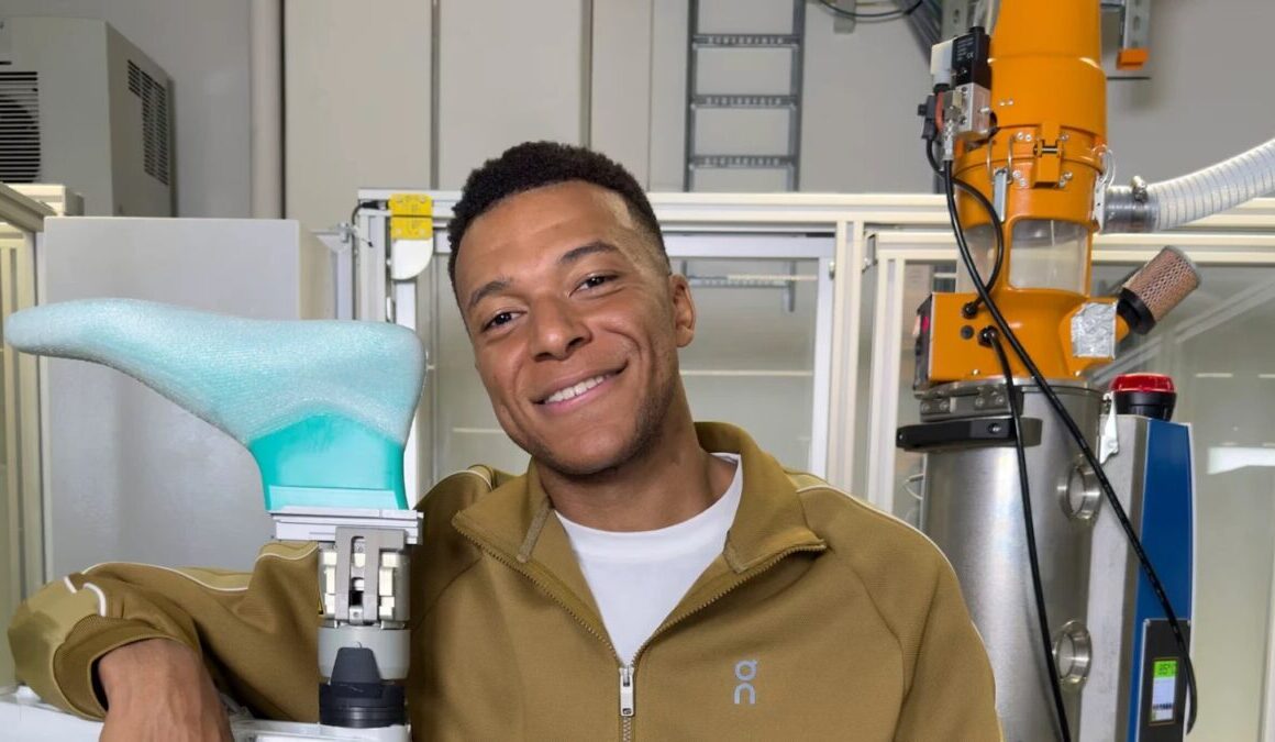 Kylian Mbappé in a tan On tracksuit resting an arm on a robotic arm inside On's innovation lab in Zurich