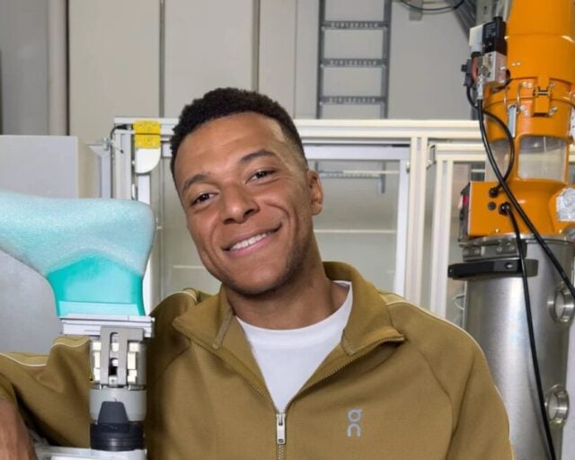 Kylian Mbappé in a tan On tracksuit resting an arm on a robotic arm inside On's innovation lab in Zurich