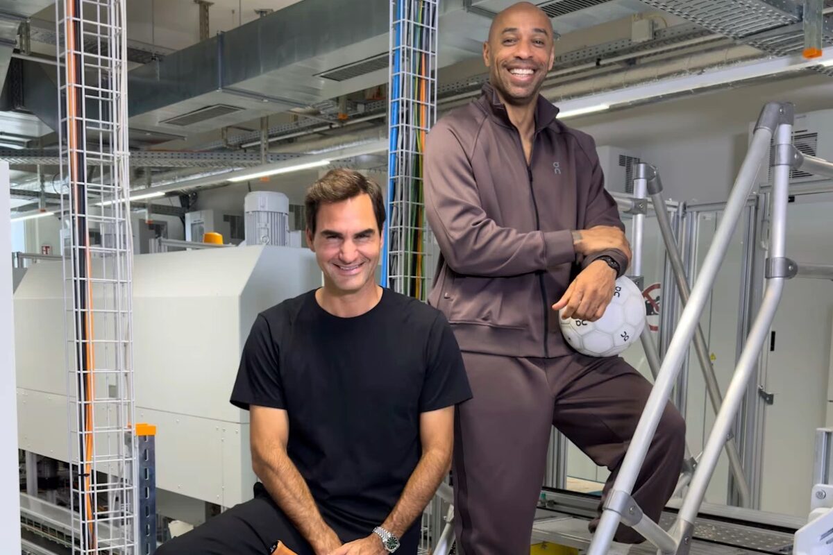 Roger Federer seated in a black T-shirt beside Thierry Henry in a brown On tracksuit holding a football, inside On's development lab