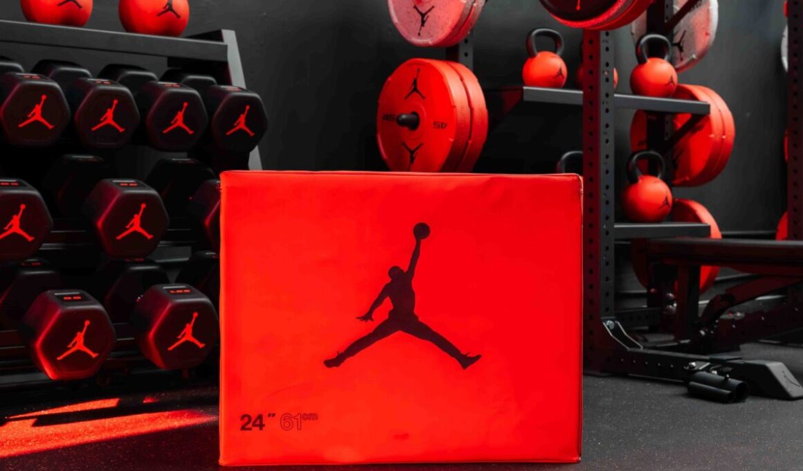 A red Jordan Strength soft plyo box with the Jumpman logo in front of black hex dumbbells, red kettlebells, bumper plates and a half rack