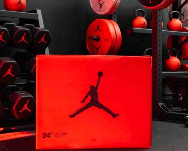 A red Jordan Strength soft plyo box with the Jumpman logo in front of black hex dumbbells, red kettlebells, bumper plates and a half rack